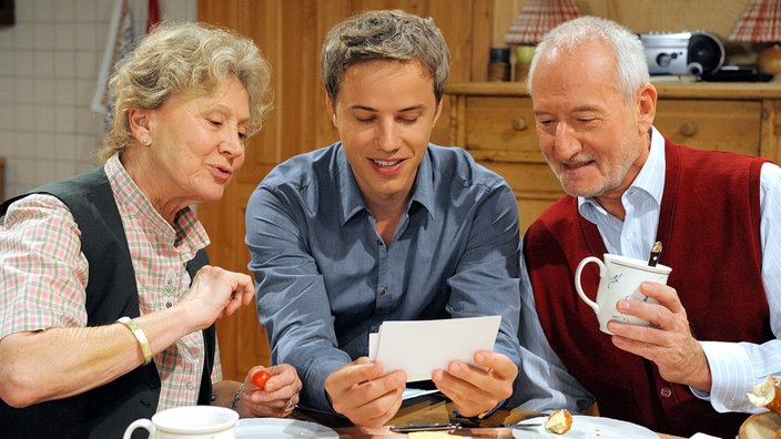 Martin (David Paryla, M.) präsentiert Hildegard (Antje Hagen, l.) und Alfons (Sepp Schauer, r.) Fotos seines Patenkindes.