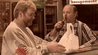 Thomas Fürst steht mit Dittsche am Tresen der Eppendorfer Grillstation. Dittsche setzt eine Flasch Burdinski Bräu an und Thomas Fürst schaut glücklich auf sein bereits von Ingo eingepacktes Essen