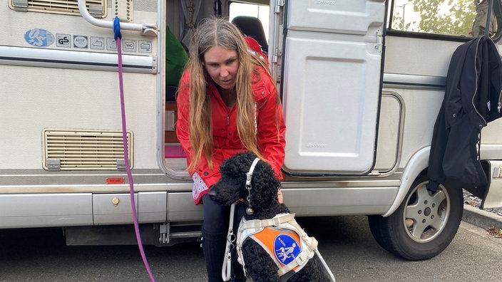 Frau mit roter Funktionsjacke streichelt Blindenhund im Hintergrund ist ein Wohnwagen zusehen