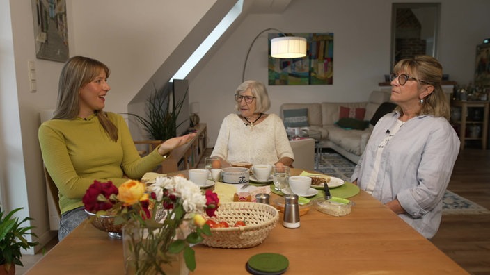 Frauen sitzen zusammen an einem mit Frühstück gedeckten Holztisch und unterhalten sich. Im Hintergrund ist das Wohnzimmer zu sehen.