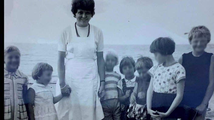 Ein altes Schwarz-Weiß-Foto zeigt ein Gruppenbild. Eine Frau in weißer Schürze steht zwischen sieben Kindern, die in einer Reihe stehen. Im Hintergrund ist das Meer zu sehen.