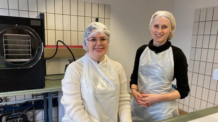 Zwei Frauen posieren in einer Werkstatt. Sie tragen Hygienekleidung