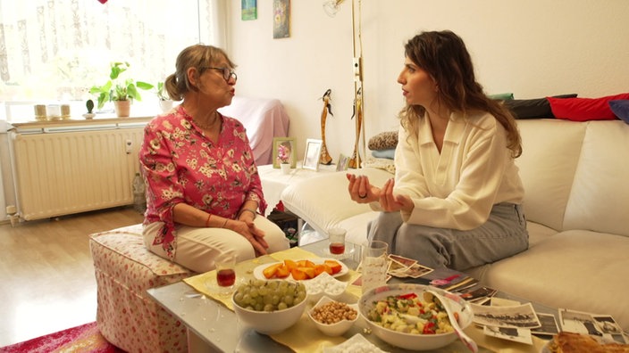 Zwei Frauen sitzen auf einer Couch im Wohnzimmer sich gegenüber und unterhalten sich miteinander