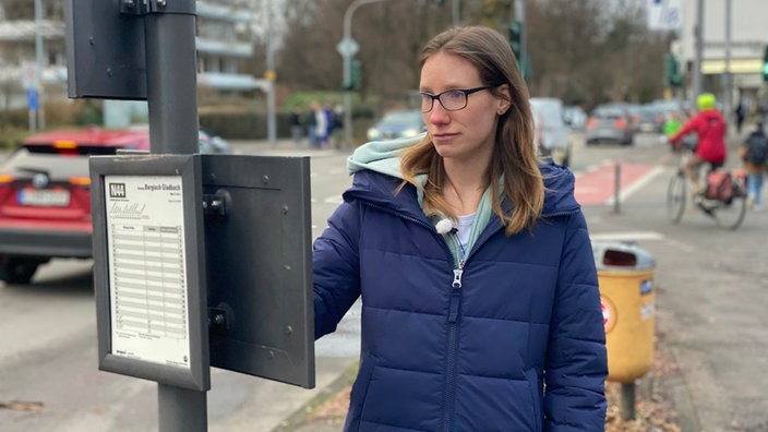 Frau steht vor einem Busfahrplan, im Hintergrund ist eine stark befahrene Straße zu sehen. Autos und eine Fahrradfahrerin sind im Bild sichtbar.