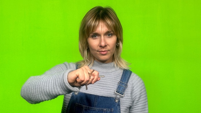 Frau steht im Greenscreen und hält ihre Faust in die Kamera. Zwischen ihren Fingern stecken einige Schlüssel