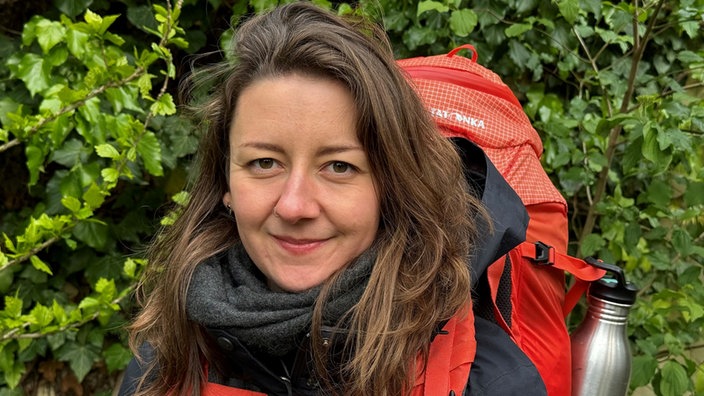 Eine Frau mit langen braunen Haaren und einem grauen Schal lächelt in die Kamera. Sie trägt einen roten Rucksack und steht vor einem Hintergrund aus grünen Blättern.