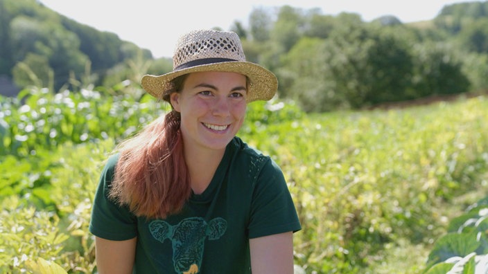 Frau mit Strohhut und grünem Tshirt steht auf grüner Wiese und lächelt in die Kamera