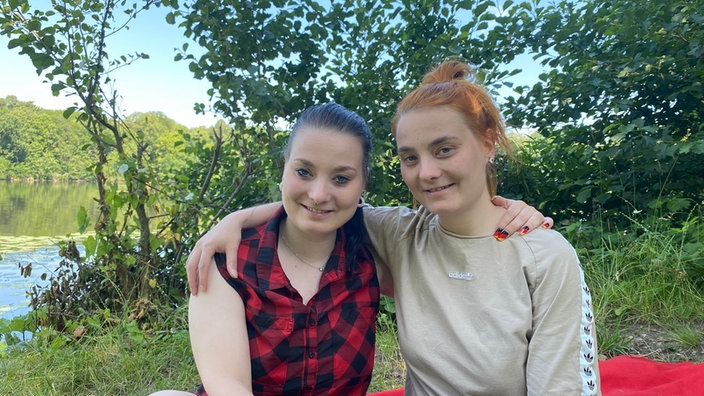 Zwillingsschwestern sitzen auf einer Bank im Wald. Im Hintergrund ist ein See zu sehen.