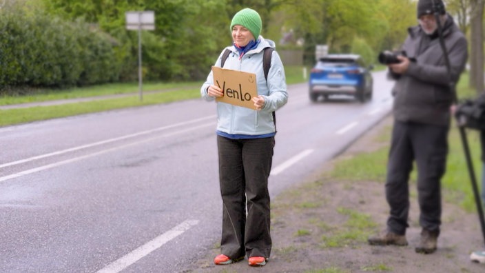 Sabine Heinrich als Anhalterin mit einem Pappschild, auf dem Venlo steht, am Straßenrand