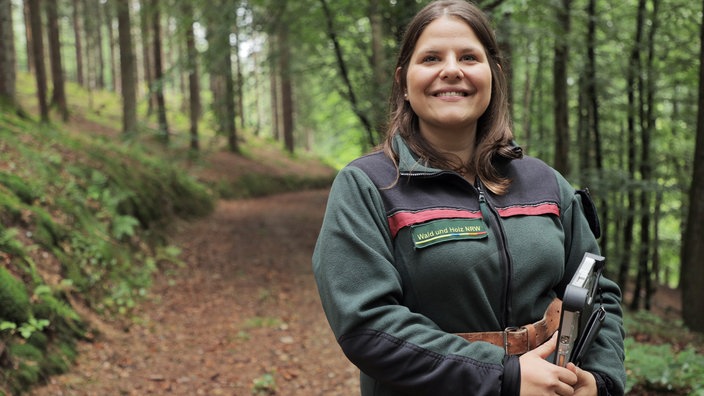 Eine junge Frau steht auf einem Waldweg und lächelt in die Kamera