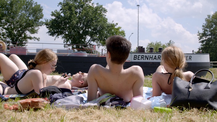 Junge Leute in Badekleidung liegen auf einer Wiese, hinter ihnen fährt ein Binneschiff vorbei