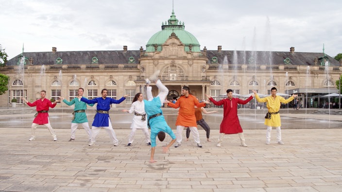 Junge Menschen in indischen Kostümen tanzen vor einem prächtigen Kurhaus