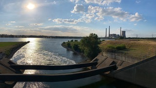 Eine Flussmündung, im Vordergrund laufen zwei Röhren über das Wasser, im Hintergrund ein Kraftwerk