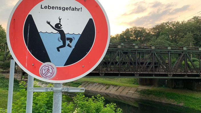 Ein Warnschild mit der Aufschrift Lebensgefahr und einer gezeichneten ertrinkenden Figur steht neben einem Fluss