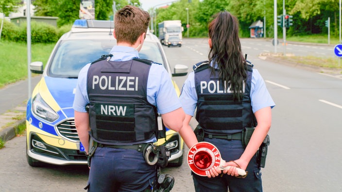 Eine Polizistin und ein Polizist stehen abwartend an einer Verkehrskontrolle neben einem Polizeiauto