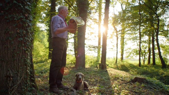 Ein Förster steht im Wald und bläst Horn, sein Hund liegt ihm zu Füßen.