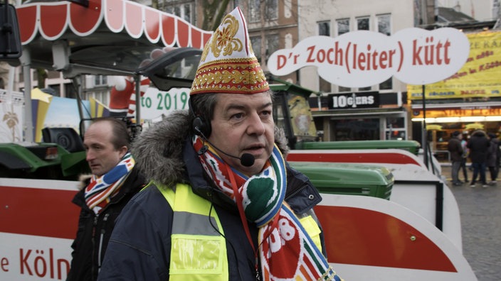 Ein Mann mit Headset, Karnevalskappe und Sicherheitsweste steht vor einem Karnevalswagen