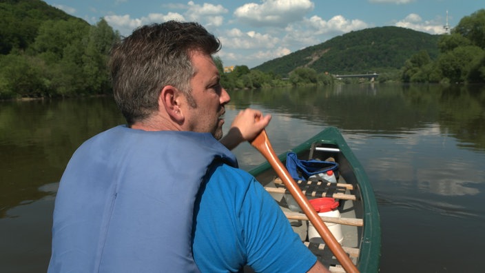 Ein Mann paddelt im Kanu durch eine grüne Vorgebirgslandschaft. 