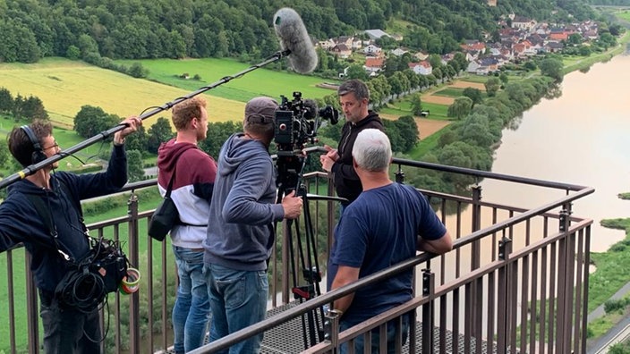 Auf einer Aussichtsplattform steht ein Kamerateam, im Hintergrund eine grüne Vorgebirgslandschaft mit Fluss. 