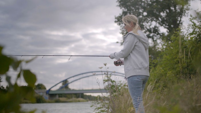 Eine Frau steht angelnd am Ufer, im Hintergrund eine Autobahnbrücke