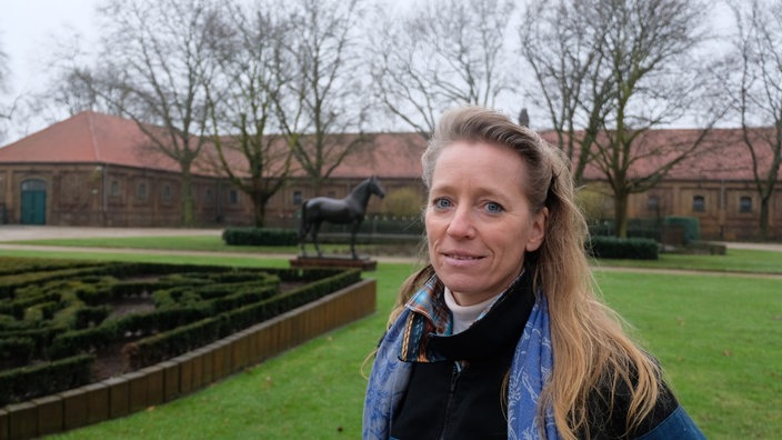 Eine Frau im Freien schaut zur Kamera, im Hintergrund eine Pferdestatue und ein Gebäude