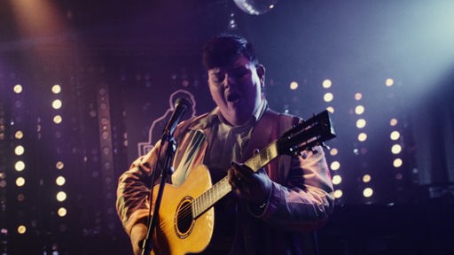 Isaak bei einem Live-Auftritt mit Gitarre auf einer Stimmungsvoll beleuchteten Bühne.