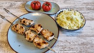 Ein Teller mit Souvlaki-Spießen, ein weiterer mit zwei Grilltomaten und ein Topf mit Krautsalat