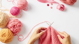 Hände stricken mit pinker Wolle auf weißem Untergrund.