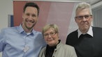 Ein junger Mann im hellblauen Hemd, eine Frau mit kurzen blonden Haaren und ein Mann mit hellem kurzem Haar posieren für ein Foto vor einer Wand mit dem Hier und heute Logo.