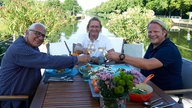 Björn Freitag und Frank Buchholz und Heinz-Dieter Fröse sitzen am Tisch und prosten sich zu. 