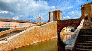 alte Backstein-Brücke mit mehreren breiten und schmalen Türmen über einem ruhigen kleinen Fluss unter blauem Himmel mit einigen Wolken, rechts und links des Wassers breite Backstein-Treppen