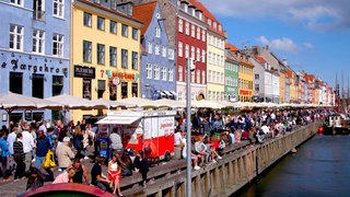 viele bunte mehrstöckige Häuser in einer Reihe unter blauem Himmel, davor weiße Sonnenschirme und viele Menschen, die entlang einer Kaimauer spazieren oder darauf sitzen, rechts Wasser und weiter hinten einige alte Segelboote