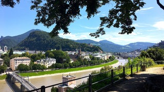 Blick von einem höher gelegenen Weg über ein Geländer im Vordergrund auf eine sonnenbeschienene Stadt mit vielen grünen Bäumen an einem schmalen Fluss, im Hintergrund grün bewachsene Hügel