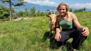 Frau mit langen, blonden Haaren, grünem Top und schwarzer Hose hockt auf einer grünen Wiese neben einer beige Ziege mit langen Hörnern, dahinter niedrige Bäume und eine sommerliche Berglandschaft