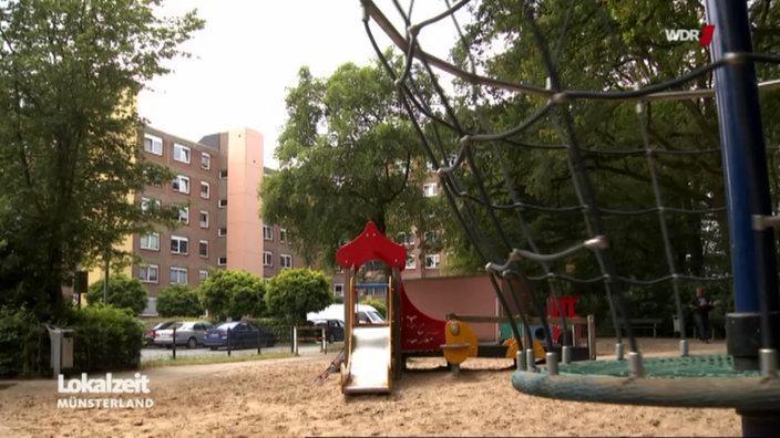 Ein von Bäumen umringter Spielplatz neben einem Mehrfamilienhaus