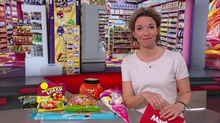 Anna Planken  steht im  Markt-Studio. Neben ihr werden verschiedene Zuckerwaren präsentiert und passend dazu im Hintergrund ein Bild eines Süßigkeitensupermarkts gezeigt.