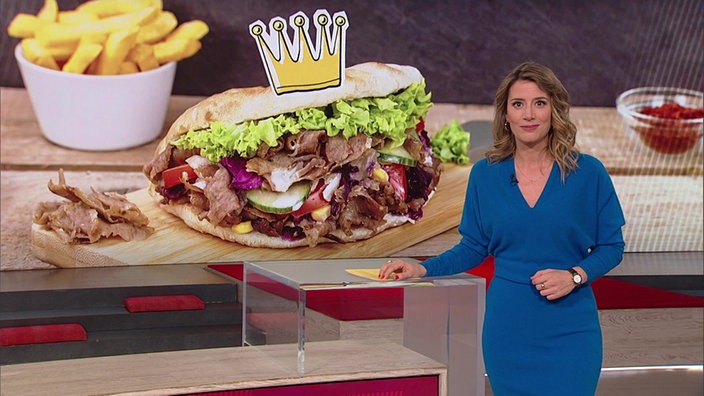 Die Moderatorin Catherine Vogel steht im Markt-Fernsehstudio. Im Hintergrund ist ein Döner und ein Becher mit Pommes zu sehen. 