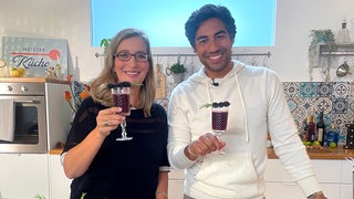 Yvonne Willicks und Nic Shanker stehen in einer Küche mit Drinks in den Händen.