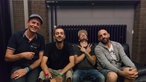 Drei Bandmitglieder mit Rockpalast-Moderator Manuel Unger im Backstage-Bereich des Summerjam Festivals. Es gibt Süssigkeiten und Cola in Plastikbechern.