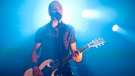 Danko Jones steht mit der Gitarre am Mikrofon und wird von hinten blau angestrahlt.