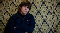 Portrait von Jake Bugg vor Wand mit üppigem Brokat. Der Musiker sieht müde und gelangweilt aus.