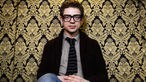 Portrait des Musikers Bernhoft, einem nicht unsympathischen Charaktergesicht mit markanter Brille, vor einem üppigen Brokathintergrund.