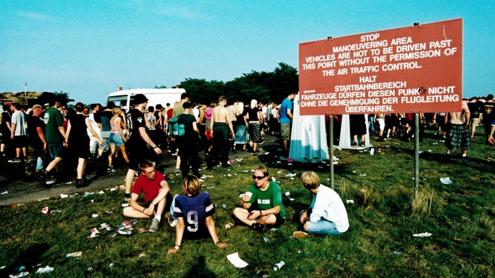 Eine Gruppe junger Festivalbesucher sitzt vor einem Schild aus dem Flughafenbetrieb: "Halt Startbahnbereich! Fahrzeuge dürfen diesen Punk (T überklebt) nicht ohne die Genehmigung der Flugleitung überfahren."