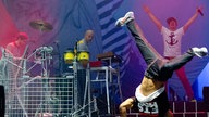 Mann macht Handstand, ein Anderer mit blauem Anker auf dem T-Shirt breitet dahinter die Arme aus.