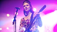 Eine Gitarristin und Sängerin Kate Nash steht an einem Mikrofon auf der Bühne und wird von weiß-lilanem Licht beleuchtet.