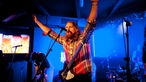 Der Sänger und Gitarrist von The Sheepdogs singt in das Mikrofon und streckt dabei beide Hände über sich in die Luft.