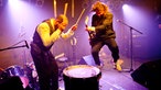 Drummer haut stehend mit dem Drumstick auf die Basedrum, der Gitarrist hüpft daneben. Im Hintergrund sieht man das zertrümmerte Schlagzeug.