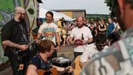 The Builders & The Butchers spielen auf dem Festivalgelände, Gitarre, Mandoline, Schellenkranz und große Tom, vorne der Kameramann, hinten die Festivalbesucher