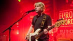 Blonder junger Mann mit Gitarre singt in ein Mikrofon im roten Bühnenlicht.