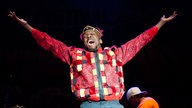 Jimmy Cliff hebt beide Arme in die Luft. Er trägt ein ausgefallen rot-kariertes Hemd und trägt eine Mütze dazu